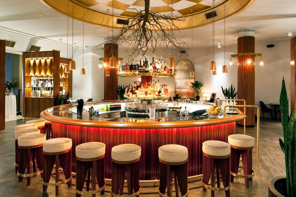 A curved hotel bar with a brass counter top, upholstered stools and hanging copper lamps