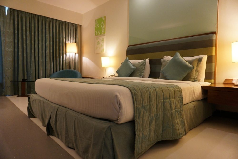 A hotel double bed with a padded headboard, bedside lamps lit and a curtained window beside it